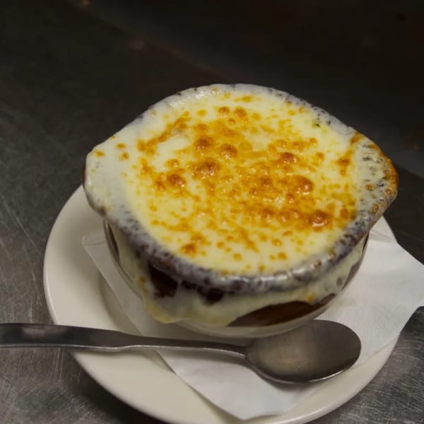 A bowl of Sommer's Place French onion soup with melted, bubbly cheese on a white plate with a spoon.